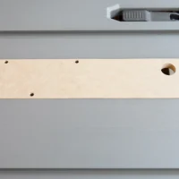Light wood MDF zero clearance insert with six mounting holes and one center opening, laid flat on a gray table saw surface next to a yellow measuring tape