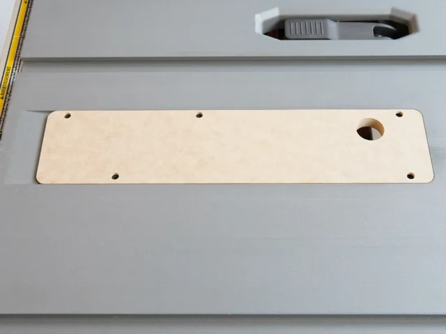 Light wood MDF zero clearance insert with six mounting holes and one center opening, laid flat on a gray table saw surface next to a yellow measuring tape