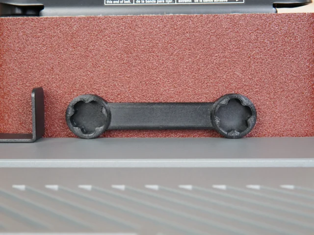 Black 3D-printed wrench with two round sockets on ends, shown above a red sanding belt on a Ridgid sander