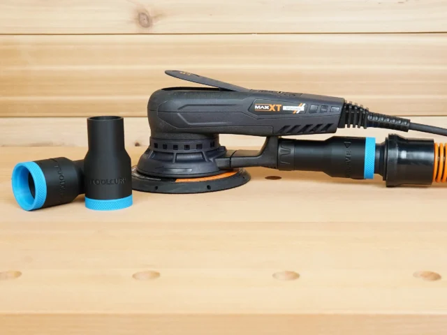 Black ToolCurve 1-1/4 inch hose adapter with blue ring lying beside a MAXXT sander, second adapter fitted to its dust port