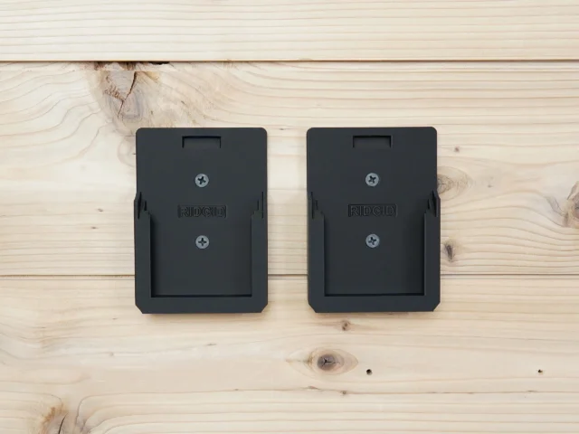 Two black Ridgid battery mounts mounted on a light wood wall, each with a recessed slot and Phillips head screws visible