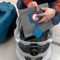 Macam reusable dust bag with blue connector held over open Makita VC4210 dust extractor, showing gray filter material and red seal mechanism