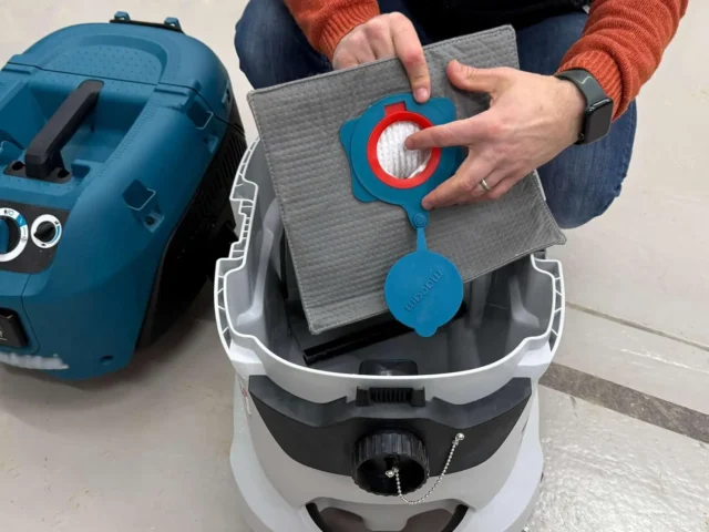 Macam reusable dust bag with blue connector held over open Makita VC4210 dust extractor, showing gray filter material and red seal mechanism