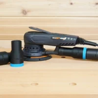 Black ToolCurve hose adapters with blue rings resting beside a MAXXT random orbital sander, one adapter joining a green extractor hose to the sander's rear port