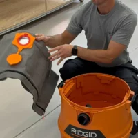 Orange Macam reusable dust bag held by a person, showing the elastic top opening and cylindrical fabric design, with an orange Ridgid shop-vac canister visible below
