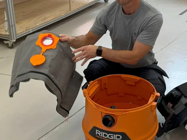 Orange Macam reusable dust bag held by a person, showing the elastic top opening and cylindrical fabric design, with an orange Ridgid shop-vac canister visible below