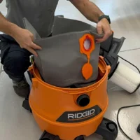 Gray polyester reusable dust bag being inserted into an orange Ridgid 12-16 gallon shop-vac, with orange elastic opening and seals visible