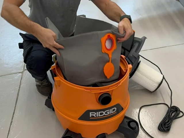 Gray polyester reusable dust bag being inserted into an orange Ridgid 12-16 gallon shop-vac, with orange elastic opening and seals visible