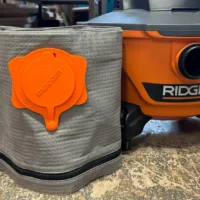 Macam reusable dust bag in tan polyester with orange plastic cap, shown next to a Ridgid shop-vac for size comparison