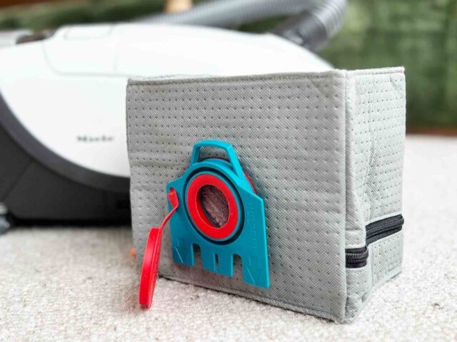 Macam reusable dust bag for Miele vacuums in light gray fabric with blue and red clip attachment, displayed next to a Miele vacuum canister