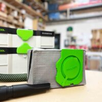 Macam reusable filter bag with gray polyester fleece body and bright green plastic head with embossed branding, displayed on workshop table with dust extractor in background