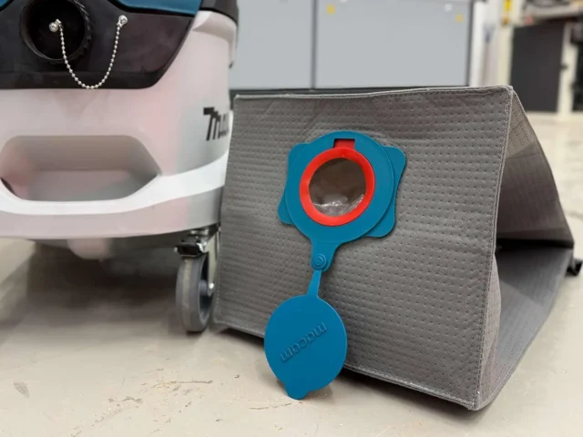 Blue Macam reusable dust bag with red ring opening, displayed in front of a gray fabric container next to a Makita dust extractor