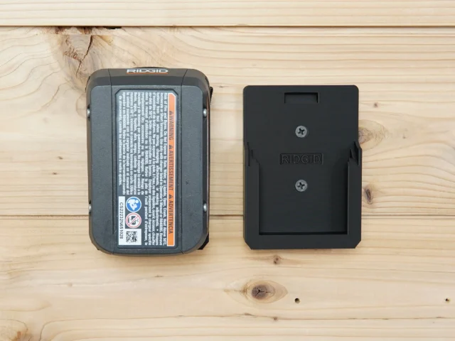 Black ToolCurve battery mount holder next to a Ridgid 18v battery on wood surface, showing the slim profile and two screw holes for wall mounting