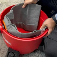 Gray polyester reusable dust bag with elastic top opening, shown pulled open inside a red Ridgid shop-vac bucket