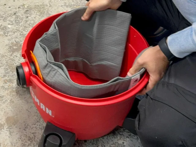 Gray polyester reusable dust bag with elastic top opening, shown pulled open inside a red Ridgid shop-vac bucket