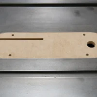Tan MDF zero clearance insert with a long slot and round opening, four mounting holes, viewed from above on a metal surface