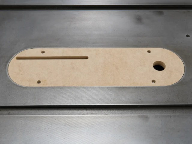 Tan MDF zero clearance insert with a long slot and round opening, four mounting holes, viewed from above on a metal surface