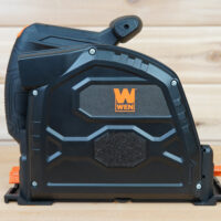 Side view of a black 7-1/4-inch track saw dust cover fitted to a WEN track saw, showing the orange WEN logo and foam pad on a wooden surface.