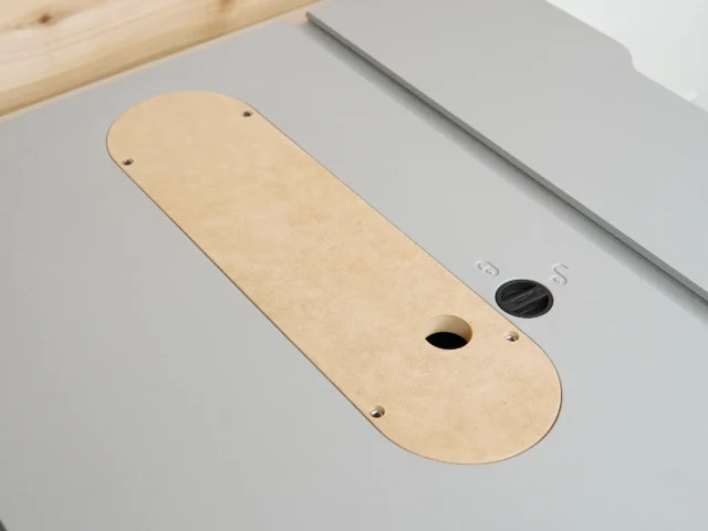 Tan MDF zero clearance insert with rounded ends, center slot, and mounting holes, positioned on a gray table saw top