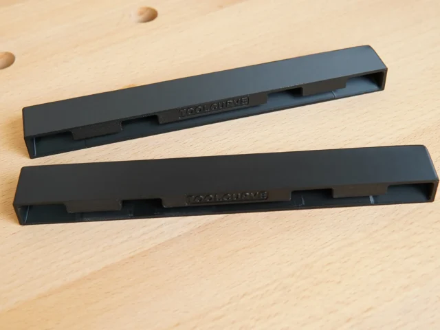 Two black PETG protective caps for PowerTec guide rails, shown separately on a wooden surface with dowel holes visible