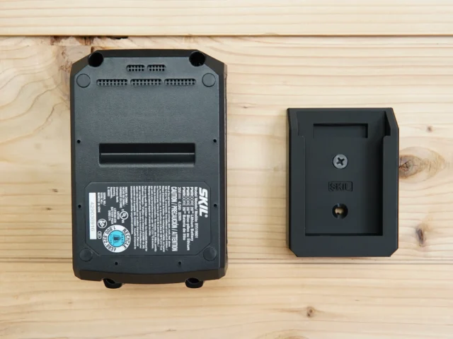 Small black wall mount with recessed slot, shown next to a SKIL 20v PWRCORE battery for size comparison