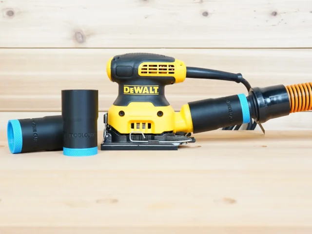 Black 1-1/4" ToolCurve hose adapter with blue ring linking a DeWalt palm sander to an orange vac hose, spare parts beside