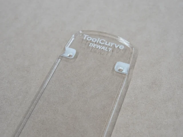 Clear acrylic zero clearance insert for Dewalt 12-inch miter saw, shown from above with two white mounting tabs visible along the sides and ToolCurve and Dewalt branding etched at the top