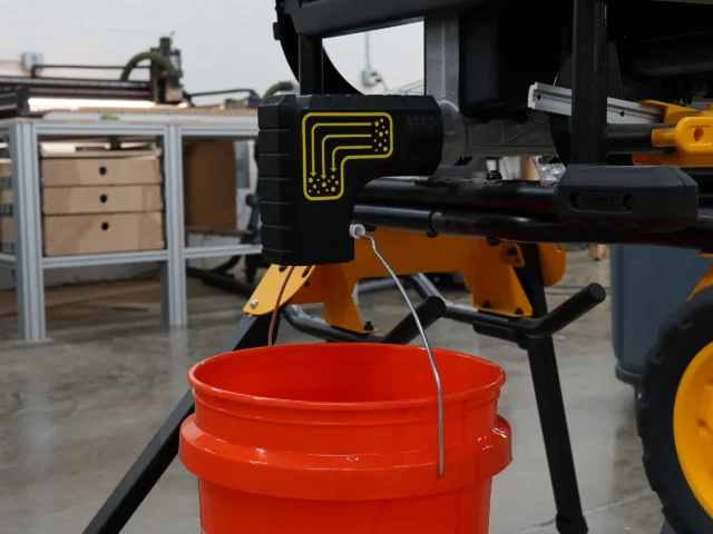 Black plastic adapter with yellow logo mounted on DeWalt table saw, directing sawdust into red bucket suspended below by wire loops
