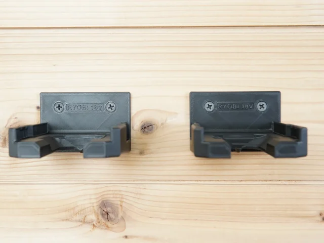 Two dark gray battery mounts for Ryobi 18v batteries mounted on light wood, showing screw holes and clip mechanisms that grip batteries from the sides