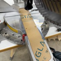 Acrylic zero-clearance router template with protective paper backing seated on a DeWalt 12-inch sliding miter saw table beside the blade