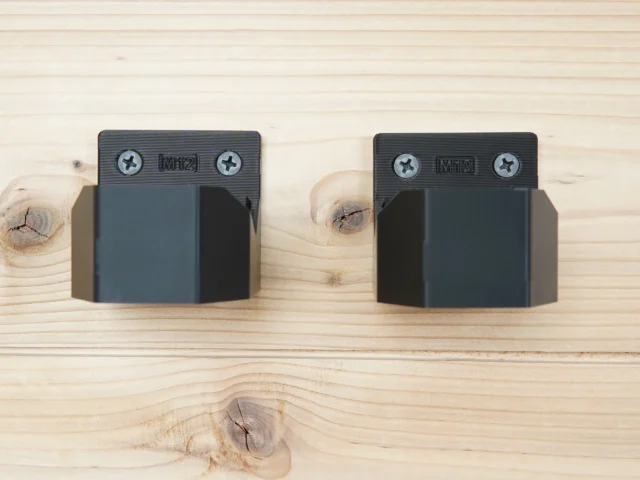 Two black battery mounts with M12 markings and Phillips screw holes mounted on light wood, showing side-profile view