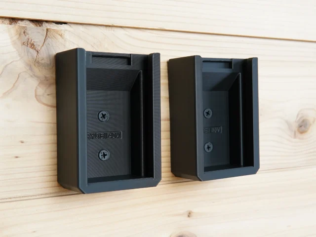 Two black plastic battery mounts mounted on wood wall, each with screw holes and internal guides sized for Ryobi 40V batteries