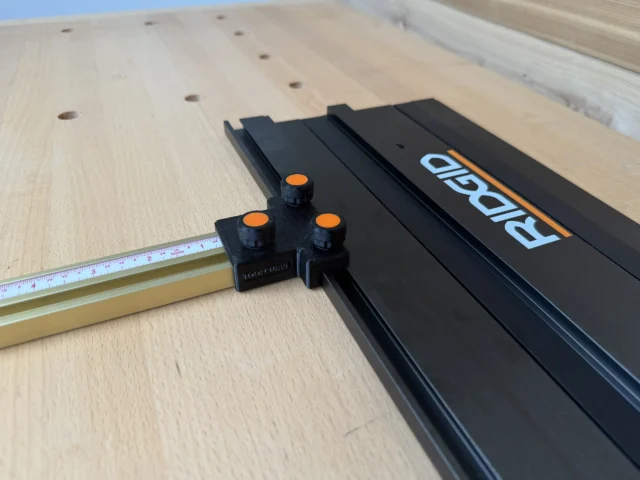 Black parallel guide adapter with orange knobs attached to Ridgid track saw rail, positioned next to tape measure on perforated workbench