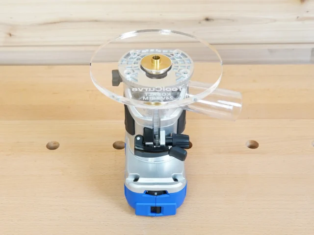 6-inch round clear acrylic router base plate mounted on blue Kobalt trim router, with brass center bushing and black mounting hardware visible
