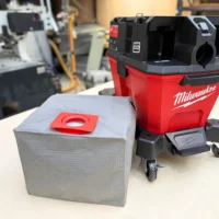 Macam reusable dust bag with red plastic collar and elastic opening, positioned next to a Milwaukee M18 Fuel Nexus vacuum on a workbench