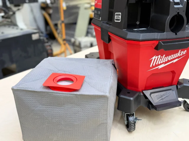 Macam reusable dust bag with red plastic collar and elastic opening, positioned next to a Milwaukee M18 Fuel Nexus vacuum on a workbench