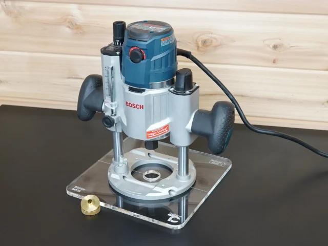 Bosch MR23EVS plunge router mounted on a 10-inch clear square acrylic guide bushing base plate, brass bushing beside it