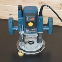 Bosch 1613EVS plunge router fitted with an 8-inch round clear acrylic base plate, brass guide bushing beside it