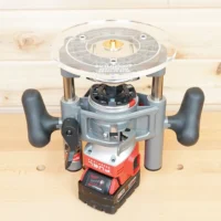 8-inch clear acrylic router base plate mounted on Milwaukee M18 2-1/4HP router, showing circular top with center bit opening and brass insert, viewed from above on workbench