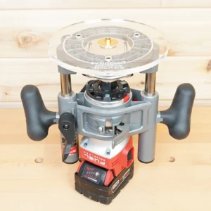 8-inch clear acrylic router base plate mounted on Milwaukee M18 2-1/4HP router, showing circular top with center bit opening and brass insert, viewed from above on workbench