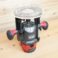 8-inch clear acrylic router base plate mounted on a red and black Skil plunge router, showing the enlarged round platform and dual handles on a wooden workbench