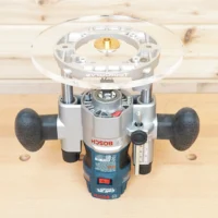 Eight-inch clear acrylic router base plate mounted on a Bosch PR011 plunge router, showing the round base from above with brass insert at center