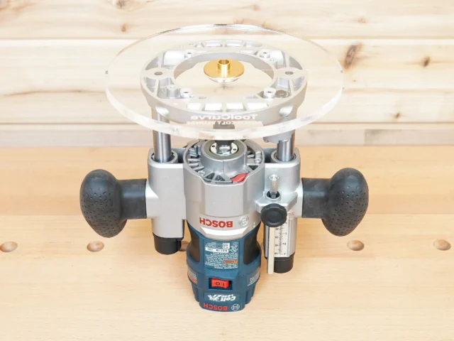 Eight-inch clear acrylic router base plate mounted on a Bosch PR011 plunge router, showing the round base from above with brass insert at center
