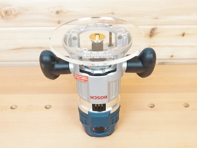 Clear acrylic router base plate with 8-inch diameter mounted on a Bosch MR23EVS plunge router, showing brass center insert and black grip handles