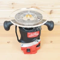 Clear acrylic 8-inch router base plate mounted on Milwaukee M18 router, showing brass bushing and guide slots, viewed from above on wooden workbench