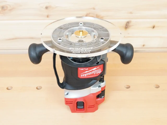 Clear acrylic 8-inch router base plate mounted on Milwaukee M18 router, showing brass bushing and guide slots, viewed from above on wooden workbench