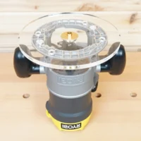 Clear acrylic 8-inch router base plate attached to Ryobi plunge router, showing brass guide bushing and black side handles on wooden workbench