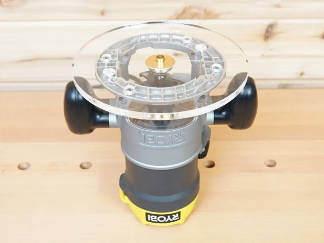 Clear acrylic 8-inch router base plate attached to Ryobi plunge router, showing brass guide bushing and black side handles on wooden workbench