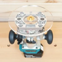8-inch clear acrylic router base plate mounted on a Makita trim plunge router, showing brass center insert and dual handles, viewed from above