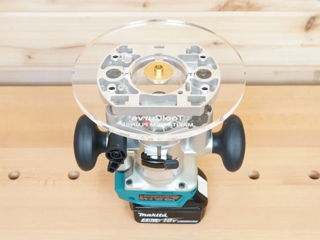 8-inch clear acrylic router base plate mounted on a Makita trim plunge router, showing brass center insert and dual handles, viewed from above